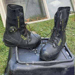 Op de afbeelding: Een paar zwarte rubberen laarzen met gele tekst die "FOLD GUSSET THIS SIDE ONLY" en "KEEP DOUBLE LACED TO HOLD FIRMLY" zegt. De laarzen staan op een zwarte plastic container.