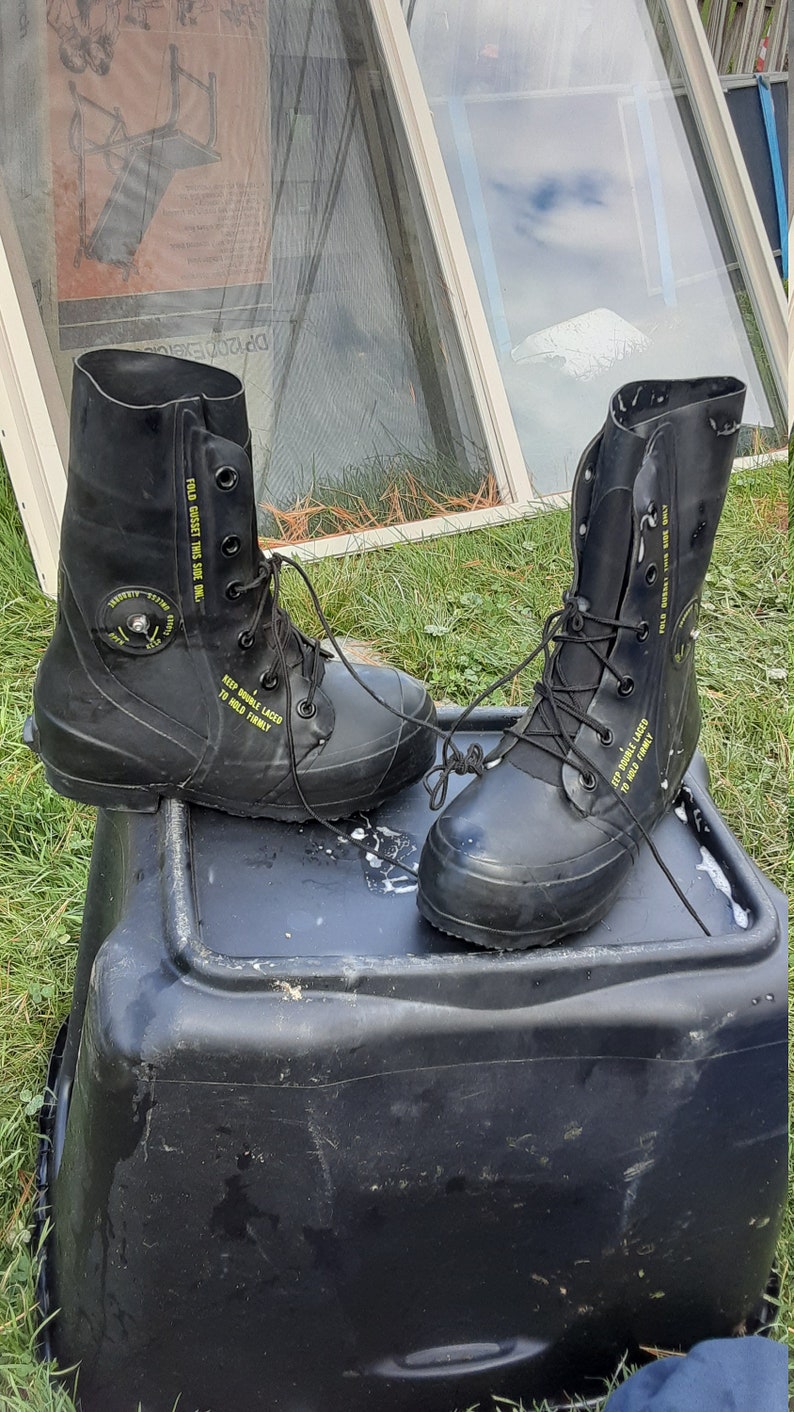 Op de afbeelding: Een paar zwarte rubberen laarzen met gele tekst die "FOLD GUSSET THIS SIDE ONLY" en "KEEP DOUBLE LACED TO HOLD FIRMLY" zegt. De laarzen staan op een zwarte plastic container.