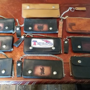 May include: Nine leather wallets with chain attachments. The wallets are black and brown with various designs and logos, including a motorcycle, a bird, and the word "Triumph".