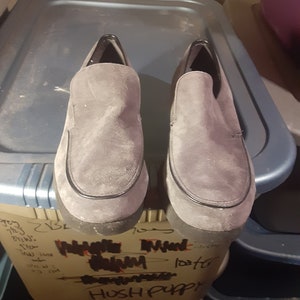 May include: A pair of gray suede Hush Puppies slip-on shoes. The shoes have a black trim around the edge and a black sole.