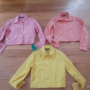 May include: Three cropped button-down shirts in shades of pink and yellow are displayed on a wooden surface. The shirts have long sleeves, collared necklines, and front pockets. The pink shirts vary in shade, while the third shirt is yellow.