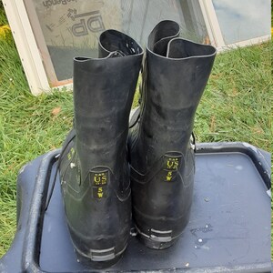 Op de afbeelding: Een paar zwarte rubberen laarzen met gele labels. De labels lezen "S.A.S. US 10-80 5 W" en "S.A.S. US 5 W".