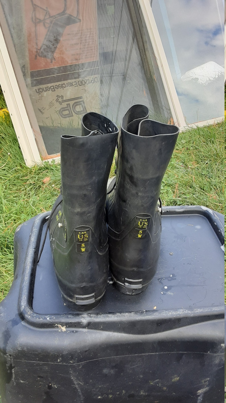Op de afbeelding: Een paar zwarte rubberen laarzen met gele labels. De labels lezen "S.A.S. US 10-80 5 W" en "S.A.S. US 5 W".