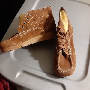May include: A pair of brown suede ankle boots with white stitching and laces. The boots have a fur lining and a rubber sole.