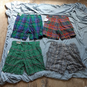 以下が含まれることがあります： 異なる色と柄の格子柄のショートパンツが4つ。ショートパンツは青い布の表面に置かれています。