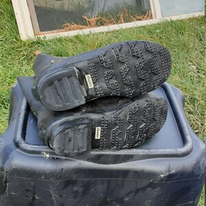 Op de afbeelding: Een paar zwarte rubberen regenlaarzen met een getextureerde zool en het woord "BATA" gedrukt op de zijkant.