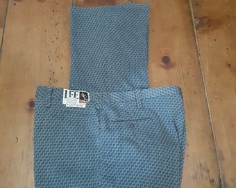 May include: A pair of gray patterned pants with a small tag that says "LEE" on the front. The pants are folded in half and laid out on a wooden floor.