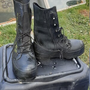 Op de afbeelding: Een paar zwarte rubberen laarzen met veters. De laarzen hebben een geel label met de tekst "TO DOUBLE LACE, HOLD FIRMLY" en "FOLD OVER, PULL FIRMLY".