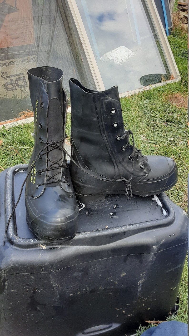 Op de afbeelding: Een paar zwarte rubberen laarzen met veters. De laarzen hebben een geel label met de tekst "TO DOUBLE LACE, HOLD FIRMLY" en "FOLD OVER, PULL FIRMLY".