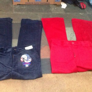 Op de afbeelding: Twee paar corduroy broeken, een in marineblauw en een in rood. De marineblauwe broek heeft een patch op het rechterbeen met een afbeelding van een persoon die een helm draagt en een vlag vasthoudt.