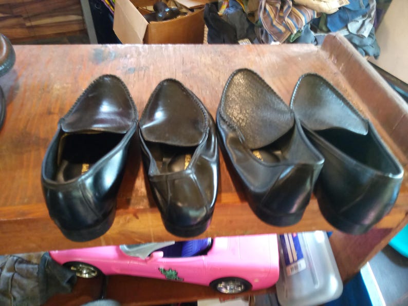 May include: Four black leather loafers with a pointed toe and a slip-on design. The loafers are arranged in a row on a wooden surface.