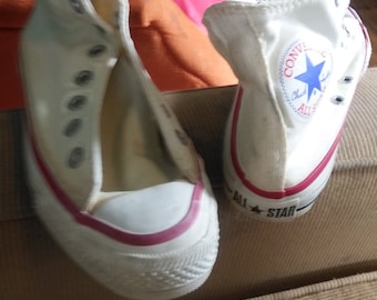 May include: A pair of white canvas high-top sneakers with red trim and a blue and white Converse logo on the side. The sneakers are sitting on a brown corduroy surface.