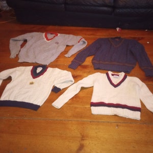 May include: Four different sweaters with v-neck collars. Two are white with navy blue and red trim, one is gray with red trim, and one is dark blue with brown trim.