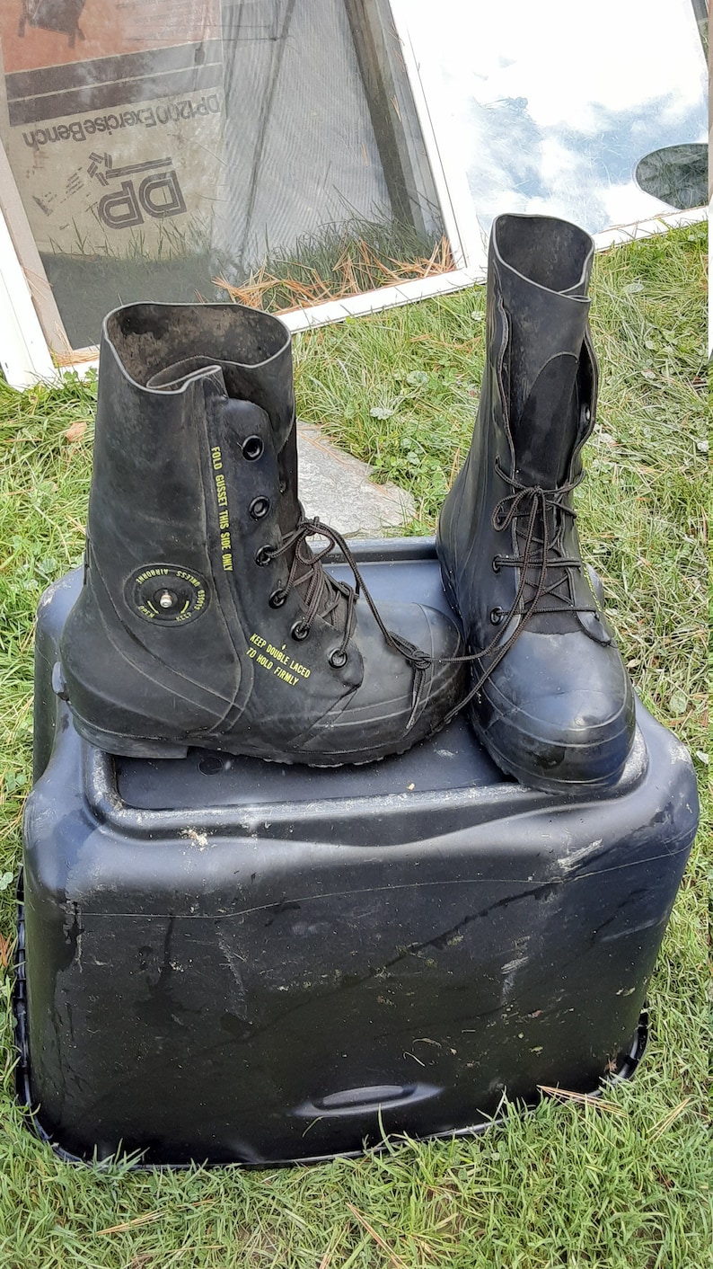 Op de afbeelding: Een paar zwarte rubberen laarzen met veters en een geel label met de tekst "FOLD GUSSET THIS SIDE ONLY" en "KEEP DOUBLE LACED TO HOLD FIRMLY". De laarzen staan op een zwarte plastic container.