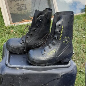 Op de afbeelding: Een paar zwarte rubberen laarzen met veters. De laarzen hebben gele tekst met de tekst "KEEP DOUBLE LACED TO HOLD FIRMLY" en "FOLD GUSSET THIS SIDE ONLY".