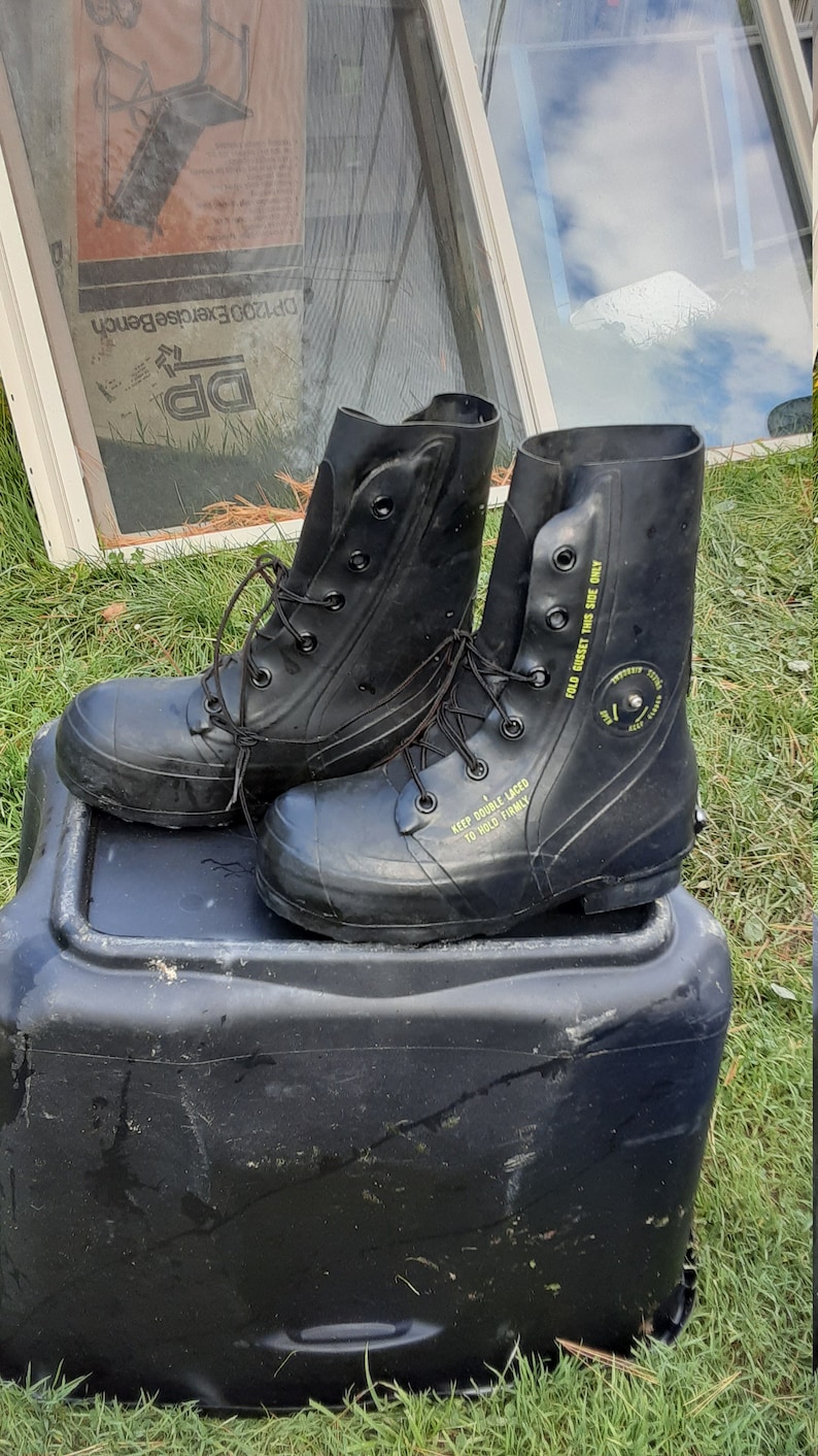 Op de afbeelding: Een paar zwarte rubberen laarzen met veters. De laarzen hebben gele tekst met de tekst "KEEP DOUBLE LACED TO HOLD FIRMLY" en "FOLD GUSSET THIS SIDE ONLY".