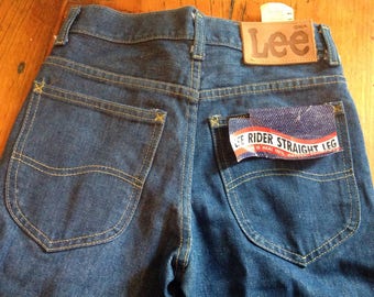 Puede incluir: Un par de pantalones vaqueros azules de mezclilla con una etiqueta de cuero marrón Lee y una etiqueta azul y blanca que dice "Lee Rider Straight Leg 100% Algodón" en los bolsillos traseros.