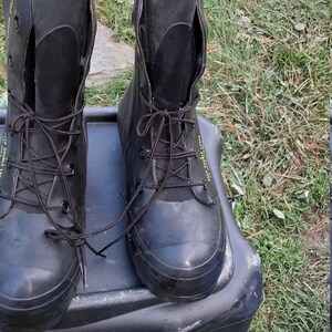 Op de afbeelding: Een paar zwarte rubberen laarzen met zwarte veters en een geel label met de tekst "Deep Double Lag" aan de zijkant. De laarzen staan op een zwarte plastic container.