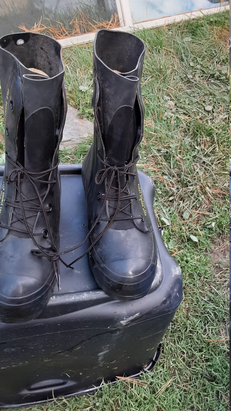 Op de afbeelding: Een paar zwarte rubberen laarzen met zwarte veters en een geel label met de tekst "Deep Double Lag" aan de zijkant. De laarzen staan op een zwarte plastic container.