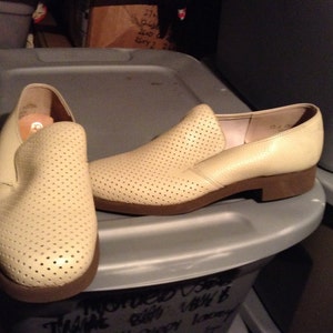 May include: Two pairs of off-white leather slip-on shoes with a perforated pattern. The shoes have a brown sole and a rounded toe.