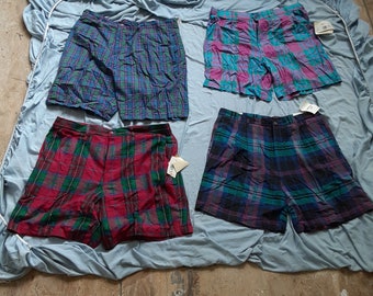 May include: Four pairs of plaid shorts in different colors and patterns. The shorts are laid out on a blue fabric surface.