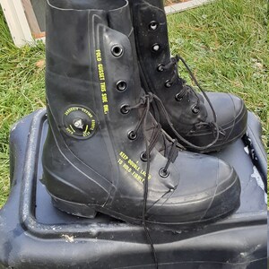 Op de afbeelding: Een paar zwarte rubberen laarzen met zwarte veters en gele tekst die "FOLD GUSSET THIS SIDE ONL." en "KEEP DOUBLE LACE D TO HOLD FIRMLY" zegt.