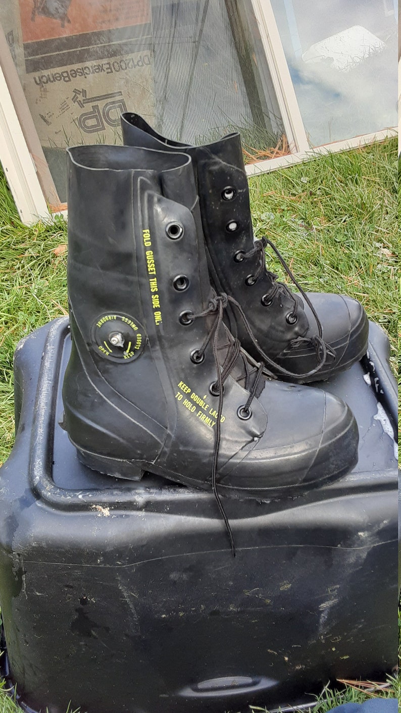 Op de afbeelding: Een paar zwarte rubberen laarzen met zwarte veters en gele tekst die "FOLD GUSSET THIS SIDE ONL." en "KEEP DOUBLE LACE D TO HOLD FIRMLY" zegt.