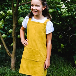 May include: A mustard yellow pinafore dress with a large front pocket. The dress is worn over a white t-shirt. The girl is smiling and standing in front of a green leafy background.