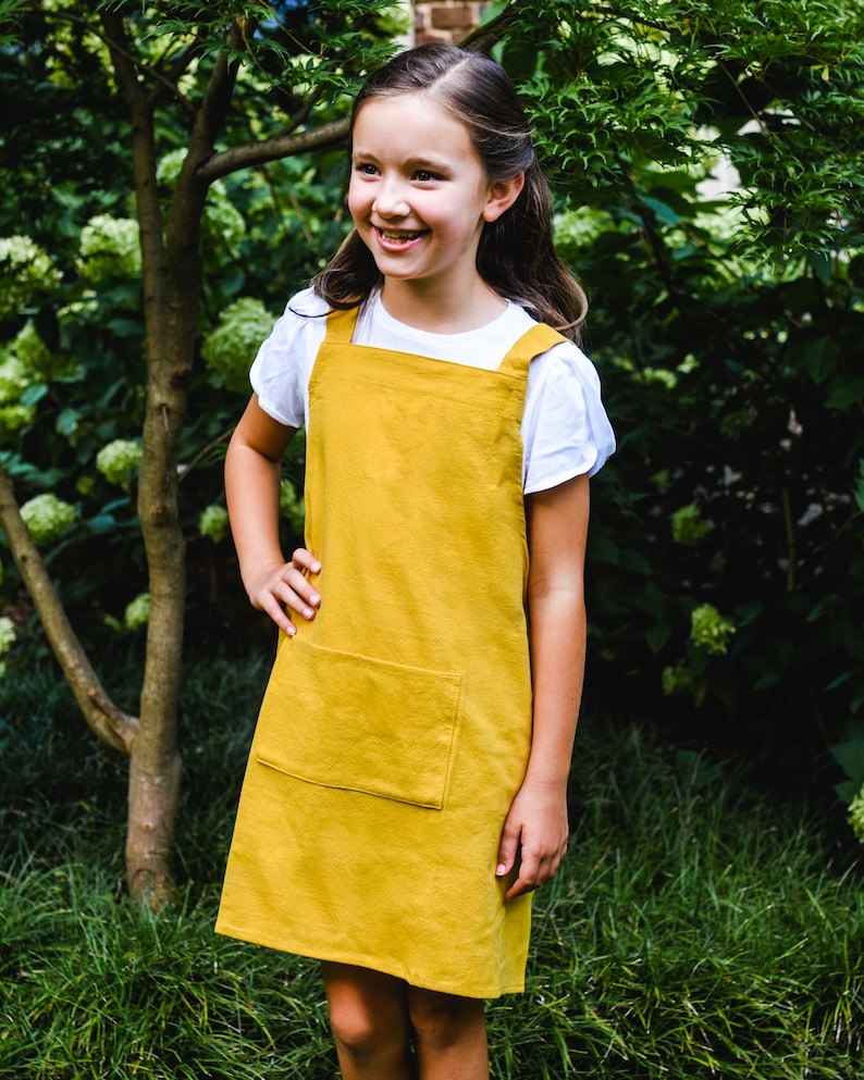 May include: A mustard yellow pinafore dress with a large front pocket. The dress is worn over a white t-shirt. The girl is smiling and standing in front of a green leafy background.