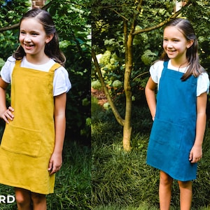 May include: Two girls wearing pinafore dresses in mustard yellow and navy blue. The dresses have a square neckline, large front pocket, and wide straps. The girls are standing in a garden setting with green foliage.