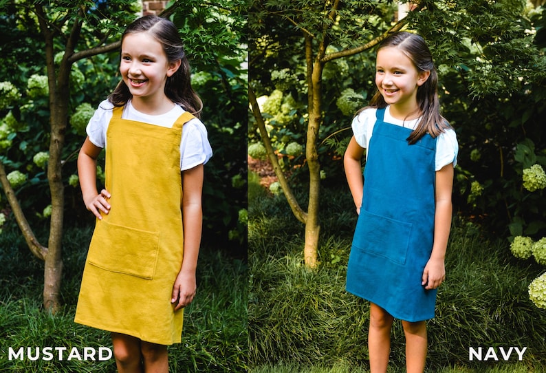 May include: Two girls wearing pinafore dresses in mustard yellow and navy blue. The dresses have a square neckline, large front pocket, and wide straps. The girls are standing in a garden setting with green foliage.