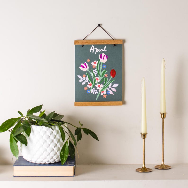 May include: A floral wall hanging with the word "April" above a bouquet of embroidered flowers in pink, red, and white. The artwork is framed with wooden bars and hangs on a white wall. Two gold candle holders with white candles are also visible.