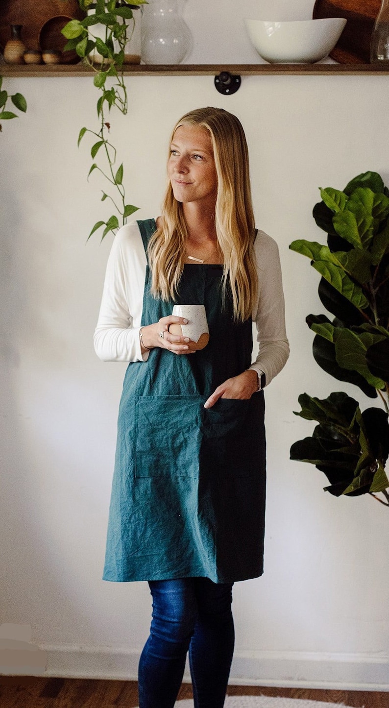 May include: A teal linen apron with a large front pocket. The apron is worn over a white long-sleeved shirt and blue jeans. A person is holding a white and tan speckled ceramic mug. The background includes greenery and a white wall.