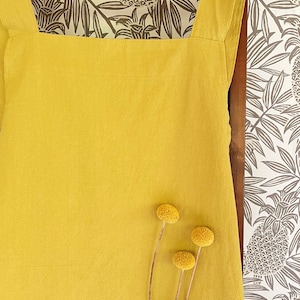 May include: Mustard yellow apron with large front pocket and wide shoulder straps. The apron is hanging on a wooden hanger against a backdrop of a patterned wallpaper with pineapple and leaf designs. Three dried yellow flowers are in front of the apron.