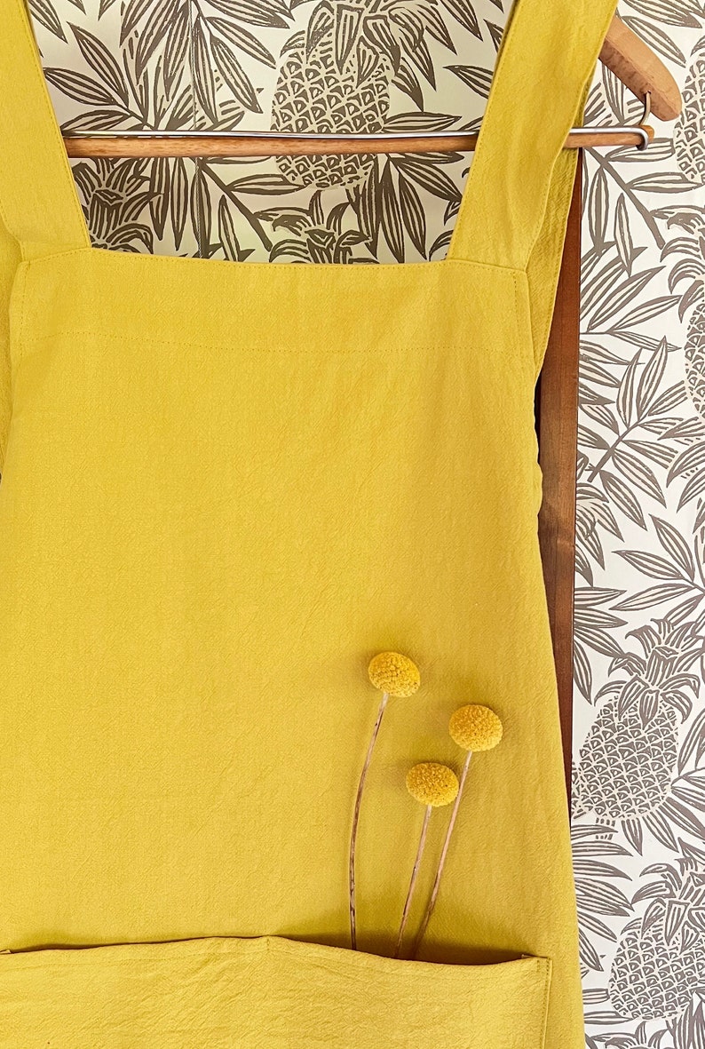 May include: Mustard yellow apron with large front pocket and wide shoulder straps. The apron is hanging on a wooden hanger against a backdrop of a patterned wallpaper with pineapple and leaf designs. Three dried yellow flowers are in front of the apron.