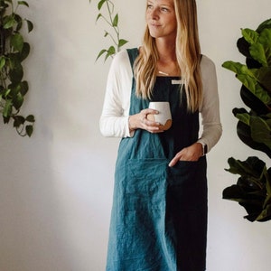 May include: A teal-colored linen apron with a square neckline and large front pockets. The apron is worn over a white long-sleeved shirt and blue jeans. The person is holding a white ceramic mug with a brown base. The background includes a shelf with plants.