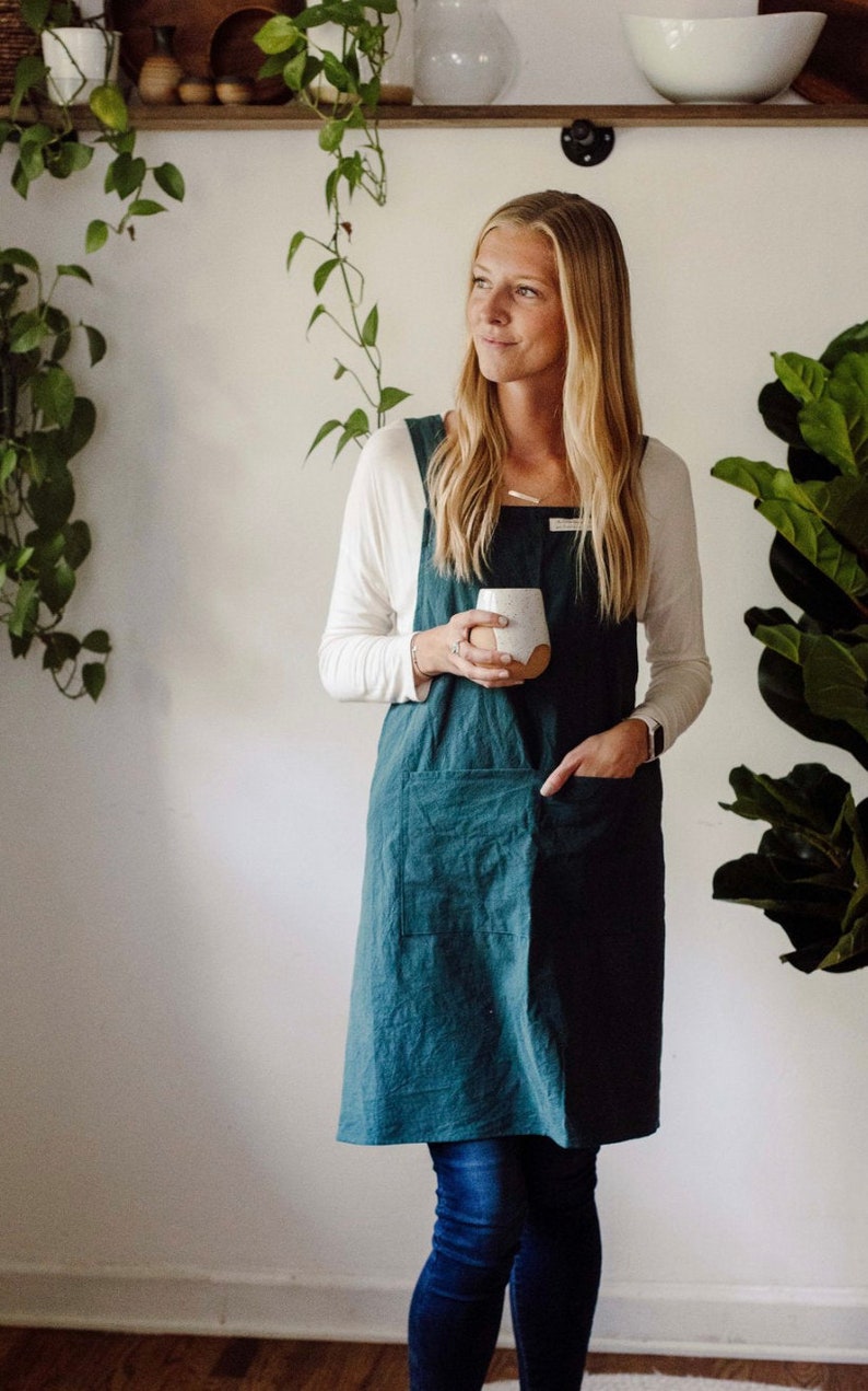 May include: A teal-colored linen apron with a square neckline and large front pockets. The apron is worn over a white long-sleeved shirt and blue jeans. The person is holding a white ceramic mug with a brown base. The background includes a shelf with plants.