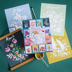 May include: A paint-by-number calendar kit with floral designs, accompanied by individual month prints. The prints feature flowers and the month's name. A paintbrush and a small container of paint are also visible.