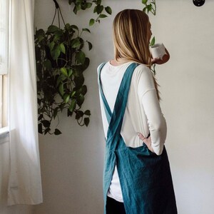 May include: A teal-colored apron with crisscross straps is worn over a white long-sleeved shirt and blue jeans. The apron is made of a textured fabric. The background includes a shelf with plants and a window with a white curtain.
