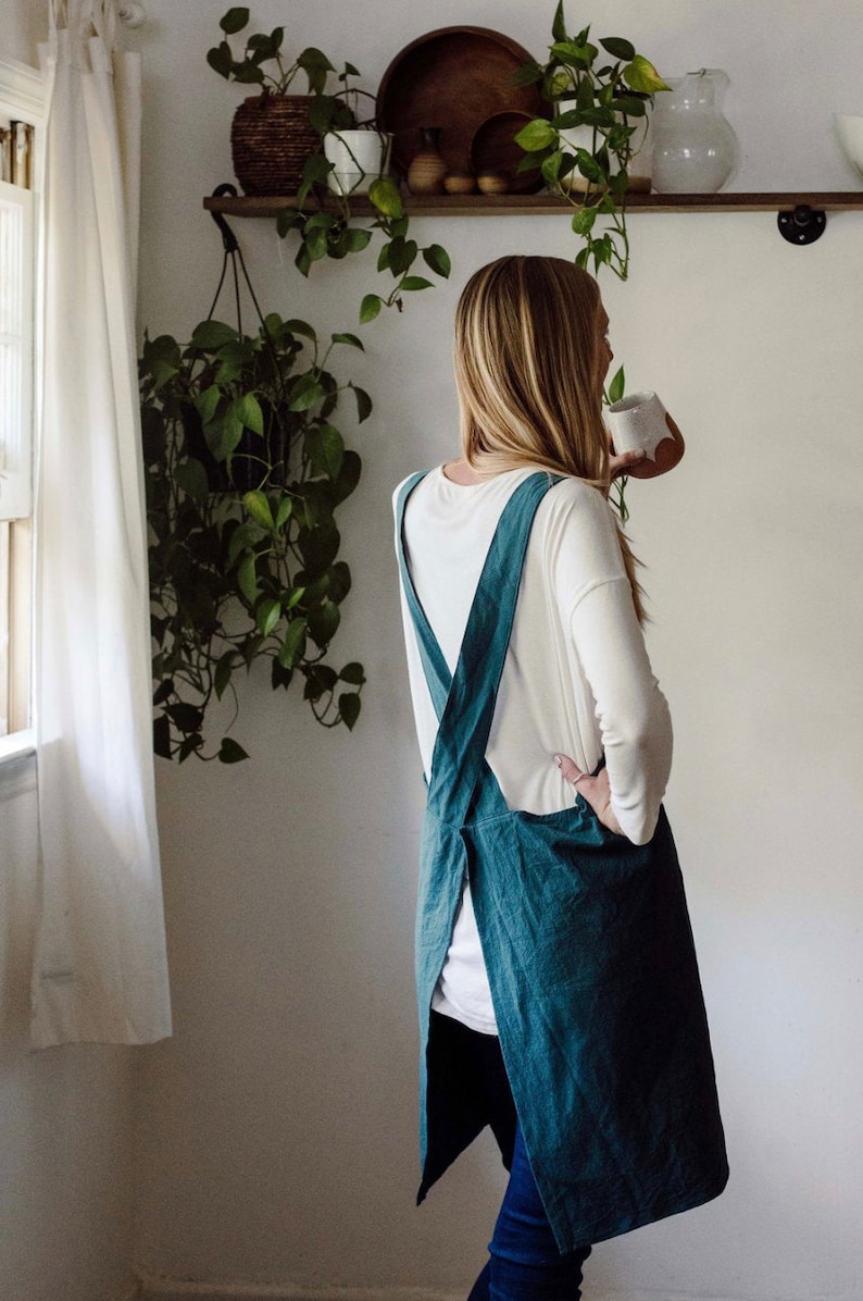 May include: A teal-colored apron with crisscross straps is worn over a white long-sleeved shirt and blue jeans. The apron is made of a textured fabric. The background includes a shelf with plants and a window with a white curtain.