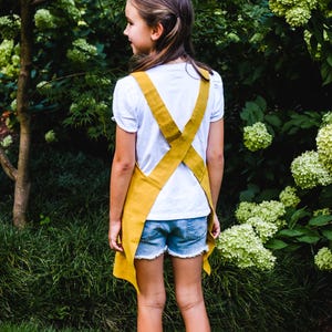 May include: A young person wearing a mustard yellow apron over a white t-shirt and denim shorts. The apron has criss-cross straps in the back. The background features green foliage and a tree.