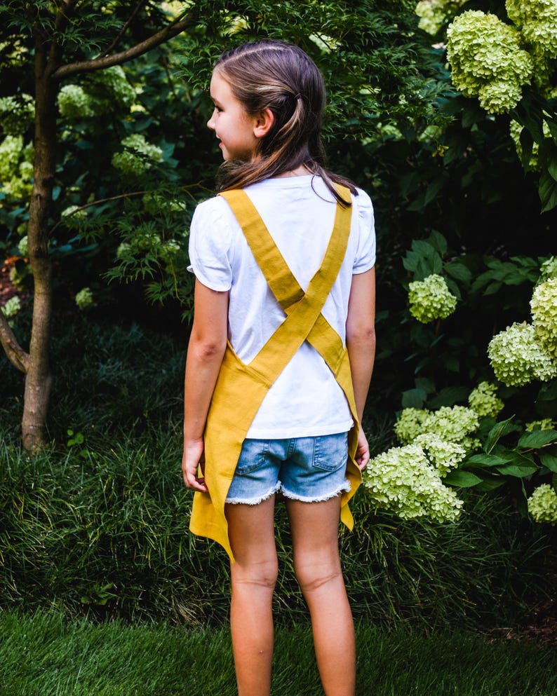 May include: A young person wearing a mustard yellow apron over a white t-shirt and denim shorts. The apron has criss-cross straps in the back. The background features green foliage and a tree.