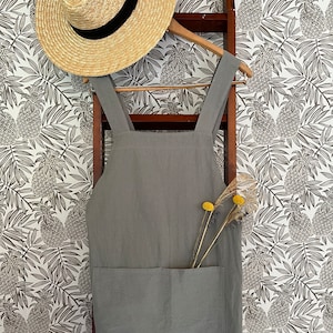 May include: A light gray linen apron with a large front pocket hangs on a wooden ladder. A straw hat with a black band rests on a wooden hanger above the apron. Dried yellow flowers are in the apron pocket. The background is a white wallpaper with a pineapple pattern.