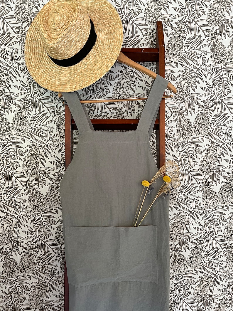 May include: A light gray linen apron with a large front pocket hangs on a wooden ladder. A straw hat with a black band rests on a wooden hanger above the apron. Dried yellow flowers are in the apron pocket. The background is a white wallpaper with a pineapple pattern.