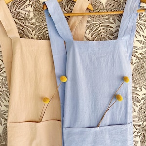 May include: Two aprons hanging on wooden hangers. One is light peach, the other is light blue. Each apron has a large front pocket and a yellow flower stem tucked inside. The background is a wallpaper with a brown pineapple pattern.