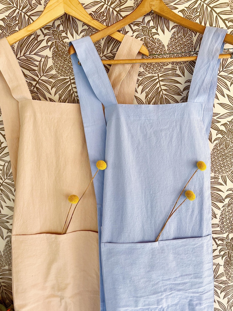 May include: Two aprons hanging on wooden hangers. One is light peach, the other is light blue. Each apron has a large front pocket and a yellow flower stem tucked inside. The background is a wallpaper with a brown pineapple pattern.