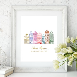 May include: A white framed print with a colorful illustration of six houses in a row. The text "Adams Morgan, WASHINGTON, DC" is printed in gold below the houses.