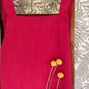 May include: A solid crimson apron with a large front pocket and cross-back straps hangs on a wooden hanger. Three yellow billy button flowers are tucked into the pocket. The background features a wallpaper with a tropical leaf pattern.