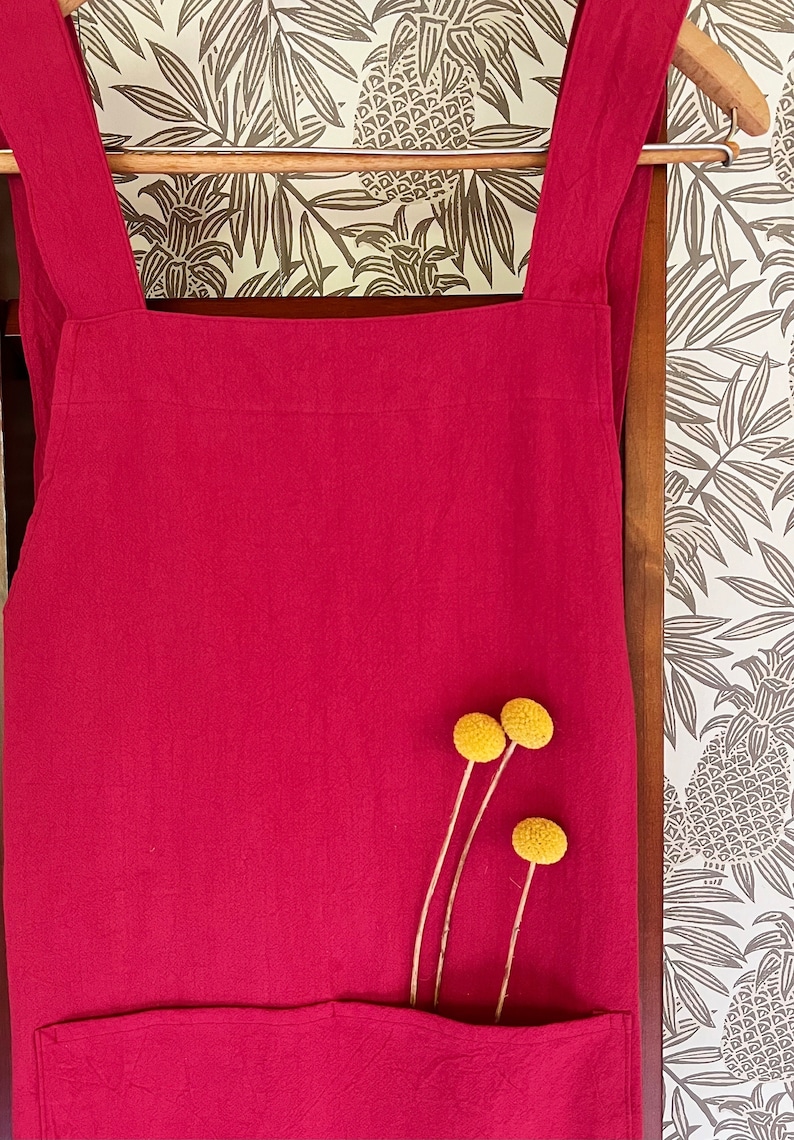 May include: A solid crimson apron with a large front pocket and cross-back straps hangs on a wooden hanger. Three yellow billy button flowers are tucked into the pocket. The background features a wallpaper with a tropical leaf pattern.