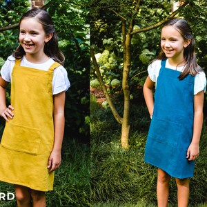 May include: Two girls wearing pinafore dresses in mustard yellow and navy blue. The dresses have a square neckline, large front pocket, and wide straps. The girls are standing in a garden setting with green foliage.